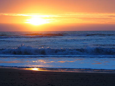 [The sun has lowered a bit and starts dipping below the cloud line above the ocean. The orb of the sun is a little less defined than earlier photos and seafoam covers most of the gold sand.]